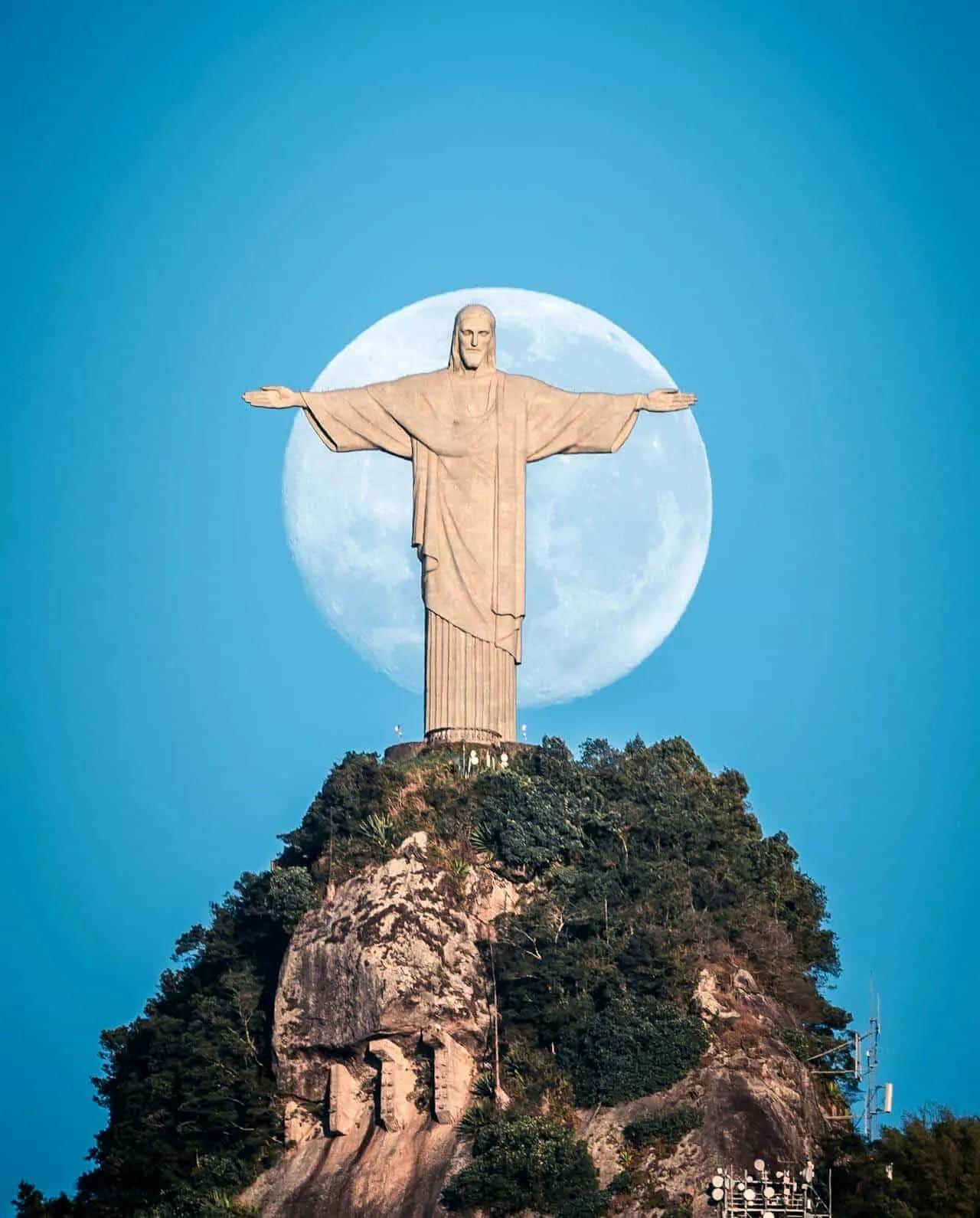 Cristo Redentor com a lua cheia ao fundo, em um céu azul claro.