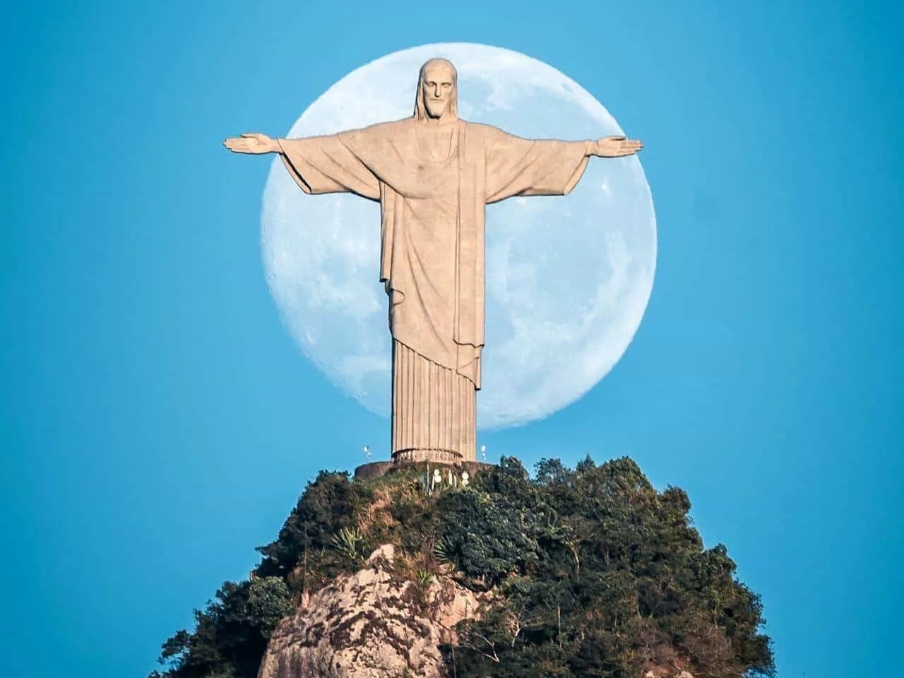Cristo Redentor com a lua cheia ao fundo, em um céu azul claro.