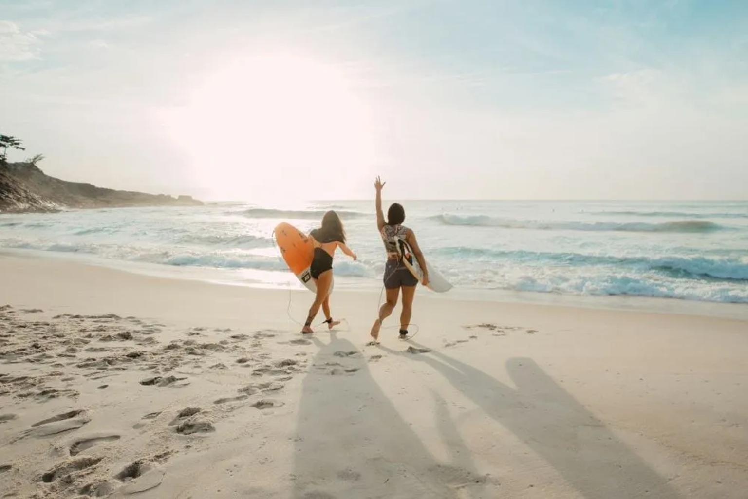 Dos surfistas caminan por la playa al atardecer, listas para disfrutar del mar.