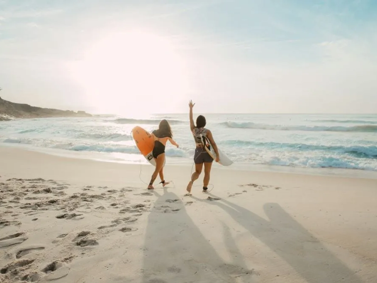 Dos surfistas caminan por la playa al atardecer, listas para disfrutar del mar.