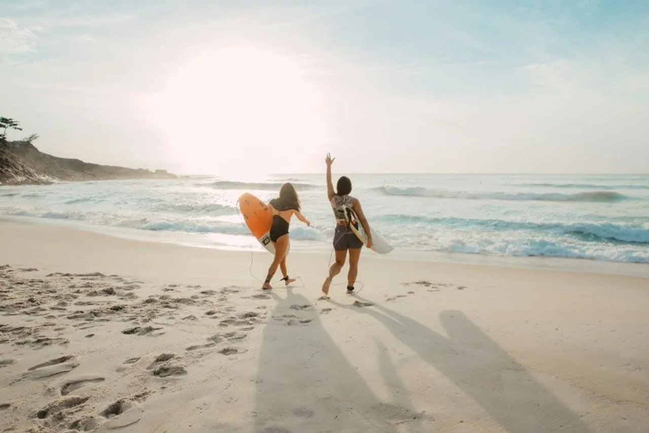 Dos surfistas caminan por la playa al atardecer, listas para disfrutar del mar.