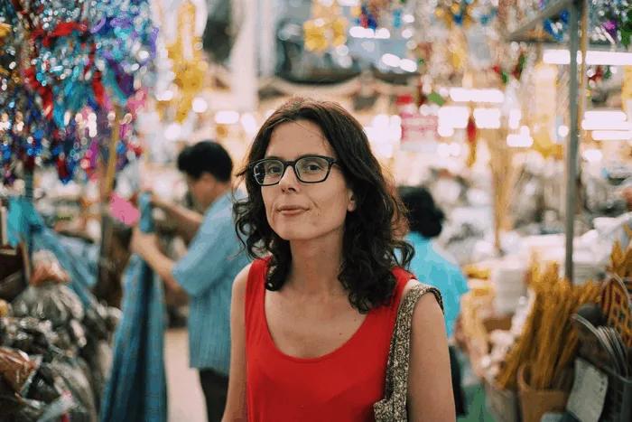 Mujer con gafas en un mercado colorido, rodeada de productos y personas.