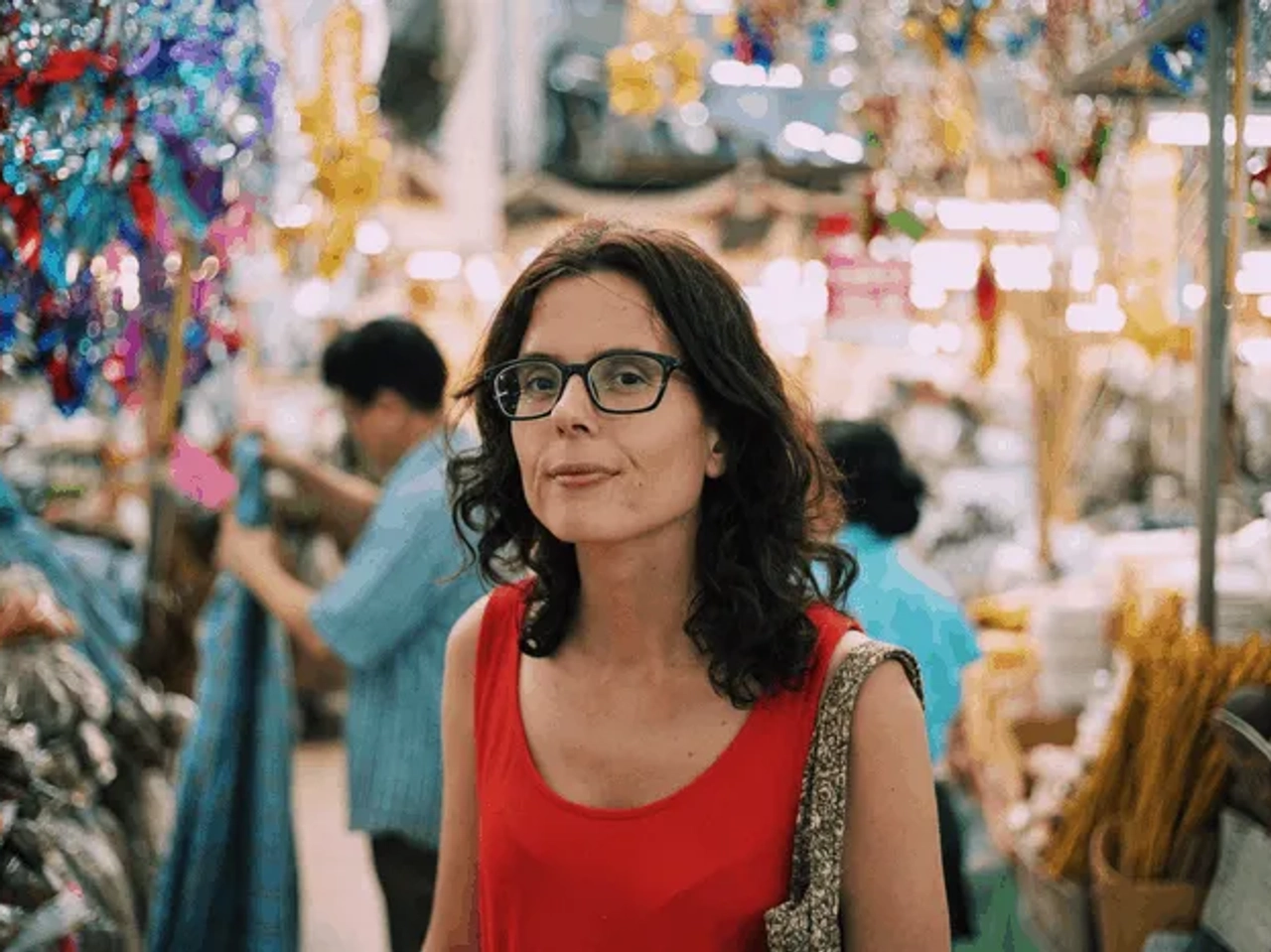 Mujer con gafas en un mercado colorido, rodeada de productos y personas.