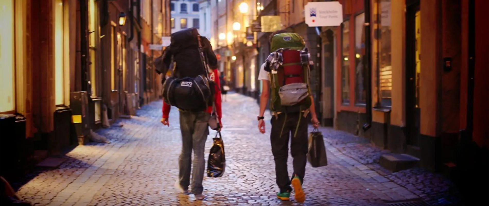Dois viajantes com mochilas caminham por uma rua de paralelepípedos iluminada.