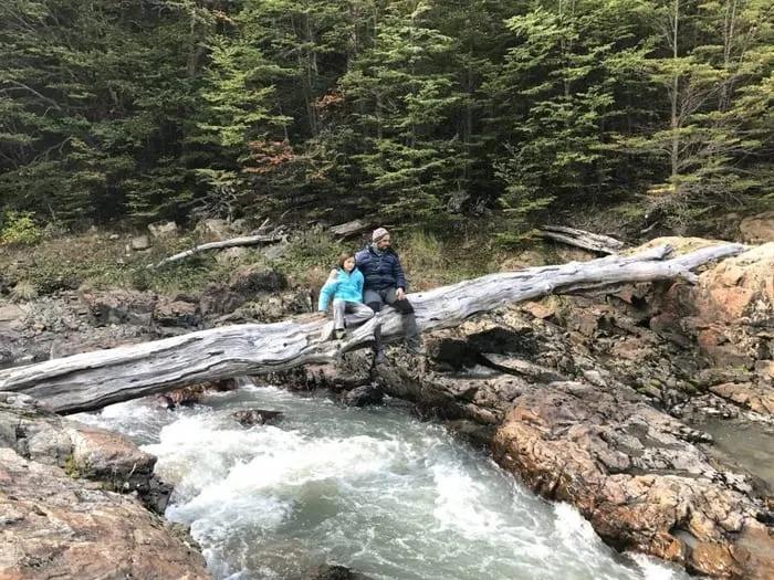 Pareja sentada sobre un tronco, junto a un río en un entorno natural.