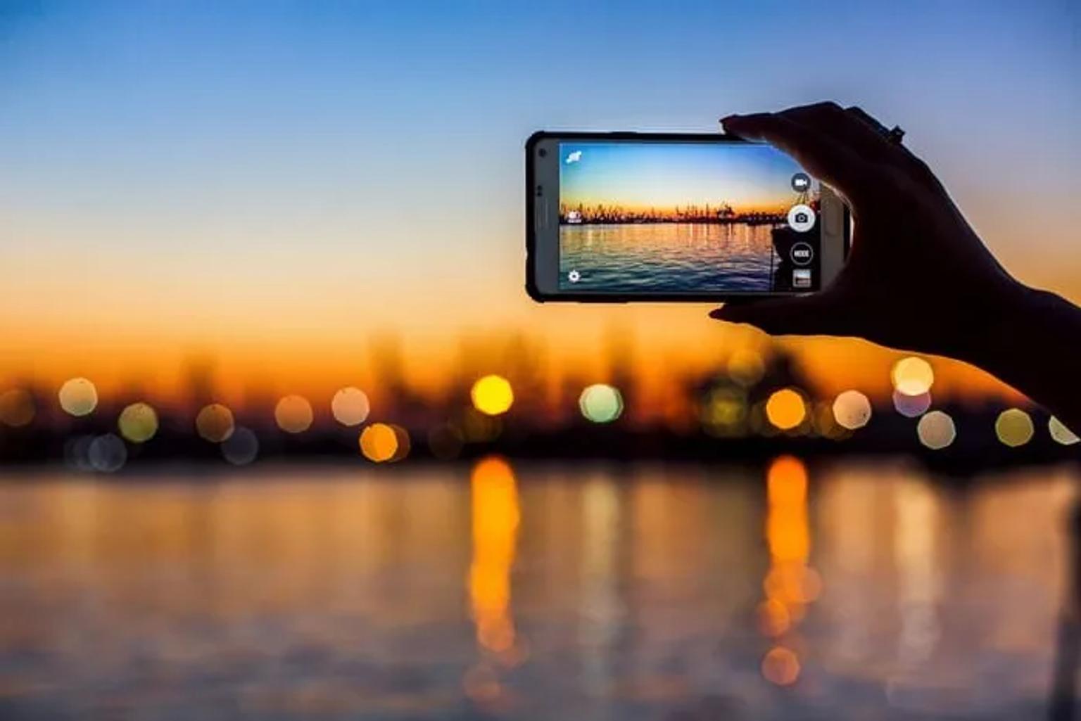 Una mano sostiene un teléfono tomando una foto de un atardecer sobre el agua.