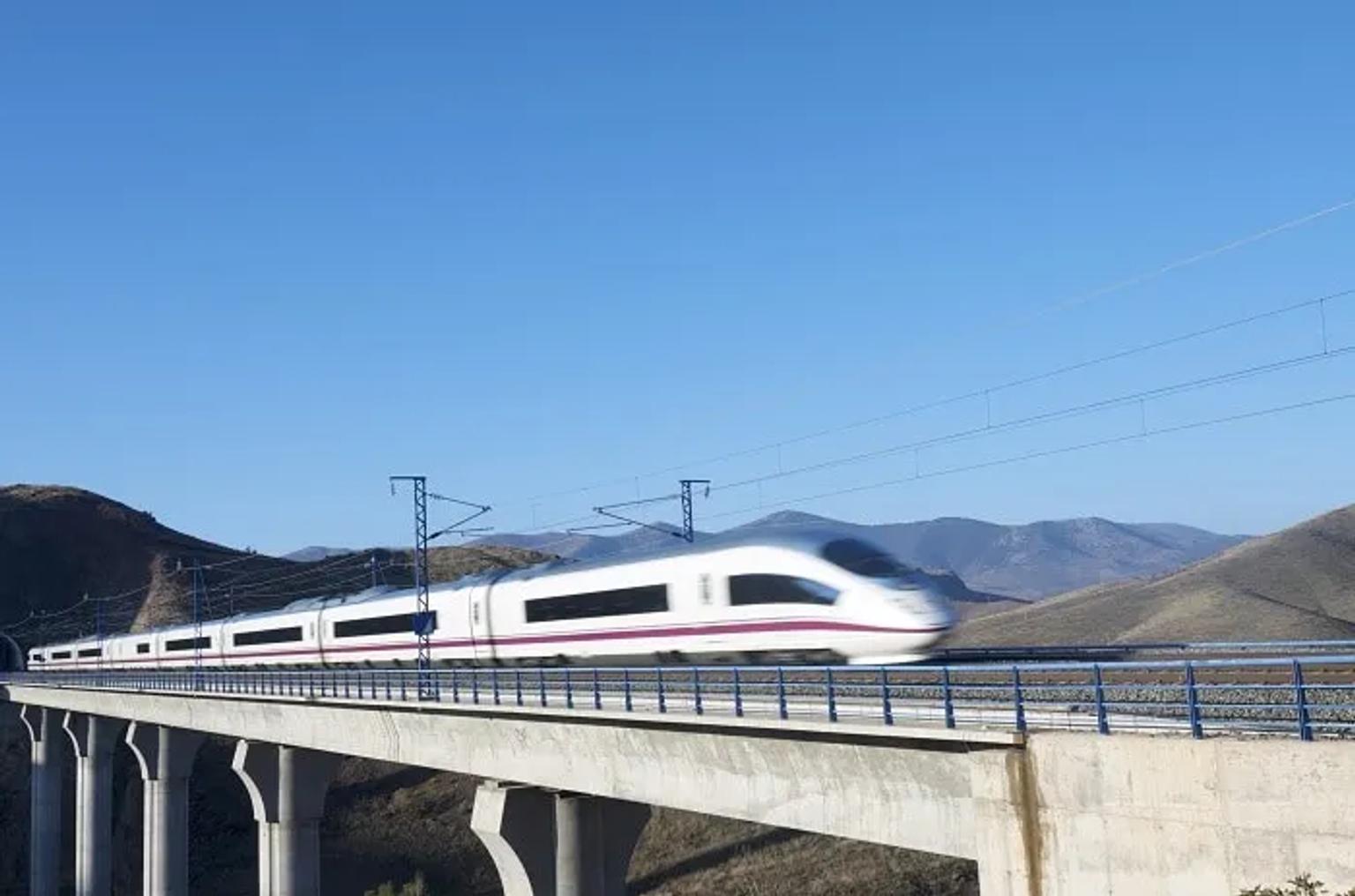 Tren AVE en movimiento sobre un puente, con paisaje montañoso al fondo.