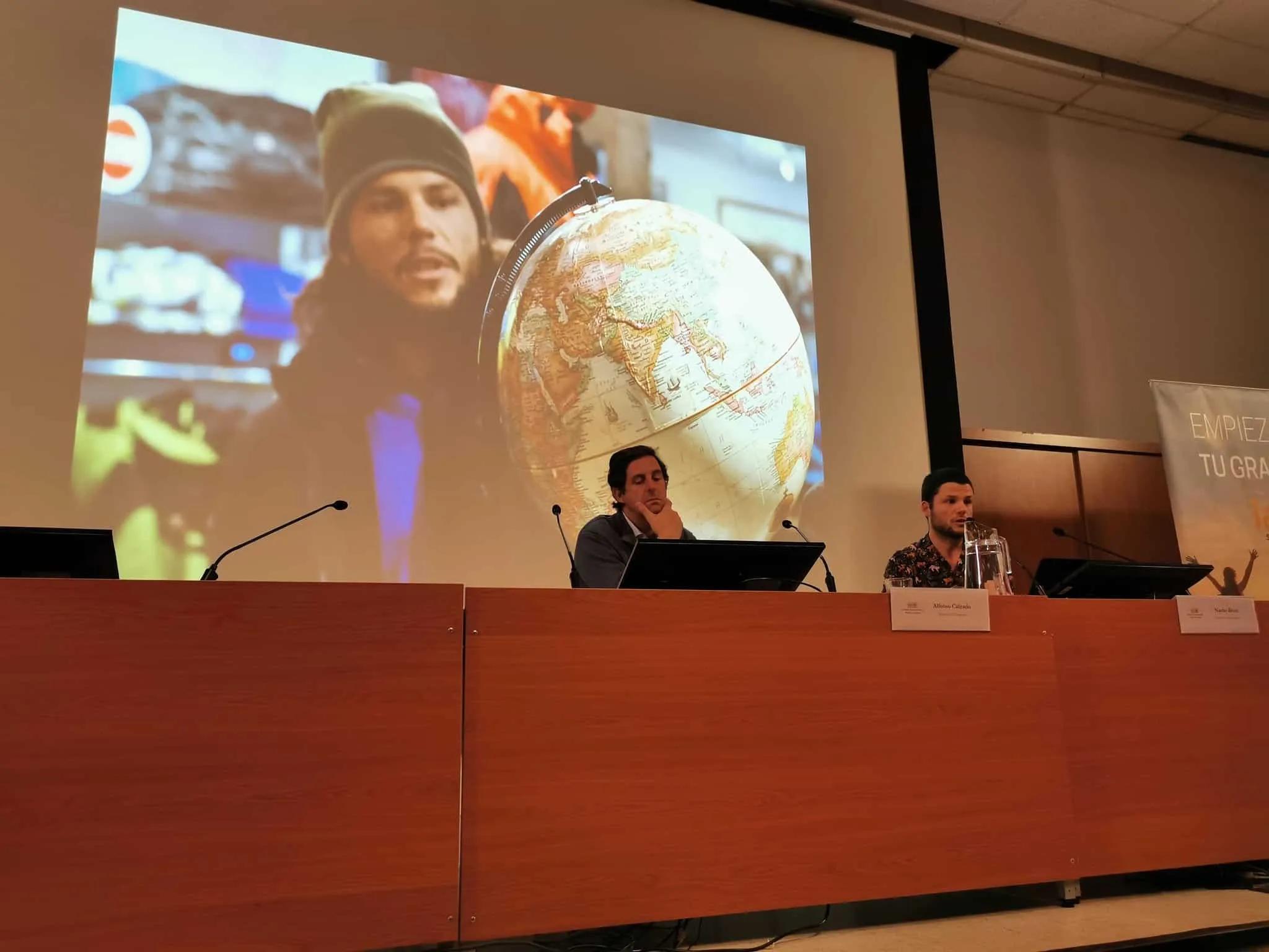 Dos ponentes discuten sobre comunicación y viajes, con un globo terráqueo de fondo.
