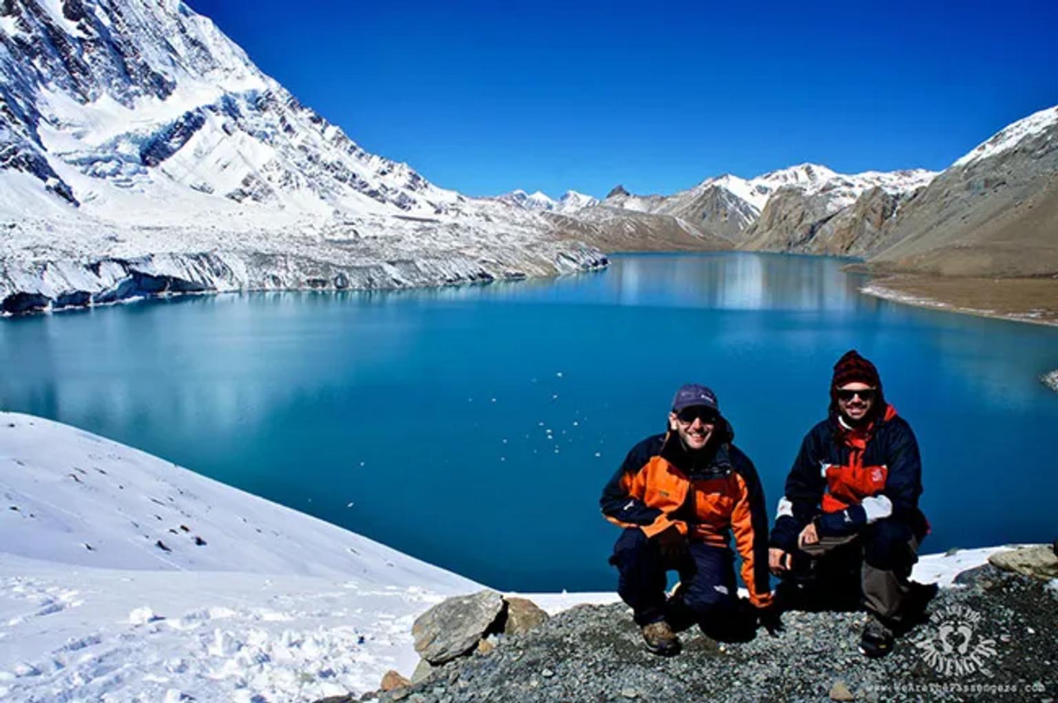 Dos personas posan frente a un lago azul rodeado de montañas nevadas.