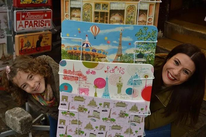 Mujer y niña sonríen detrás de postales de París en un mercado.