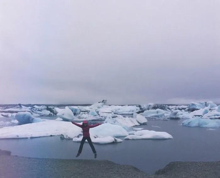 Mujer saltando frente a icebergs en un paisaje helado y nublado.