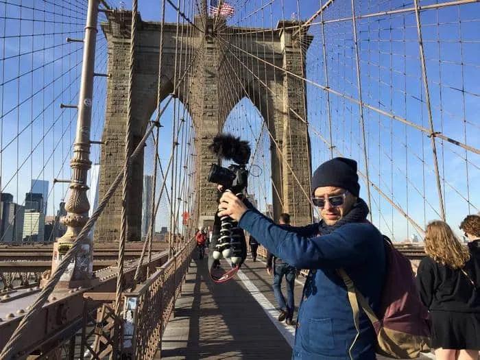 Hombre con cámara en el Puente de Brooklyn, fondo urbano y cielo despejado.