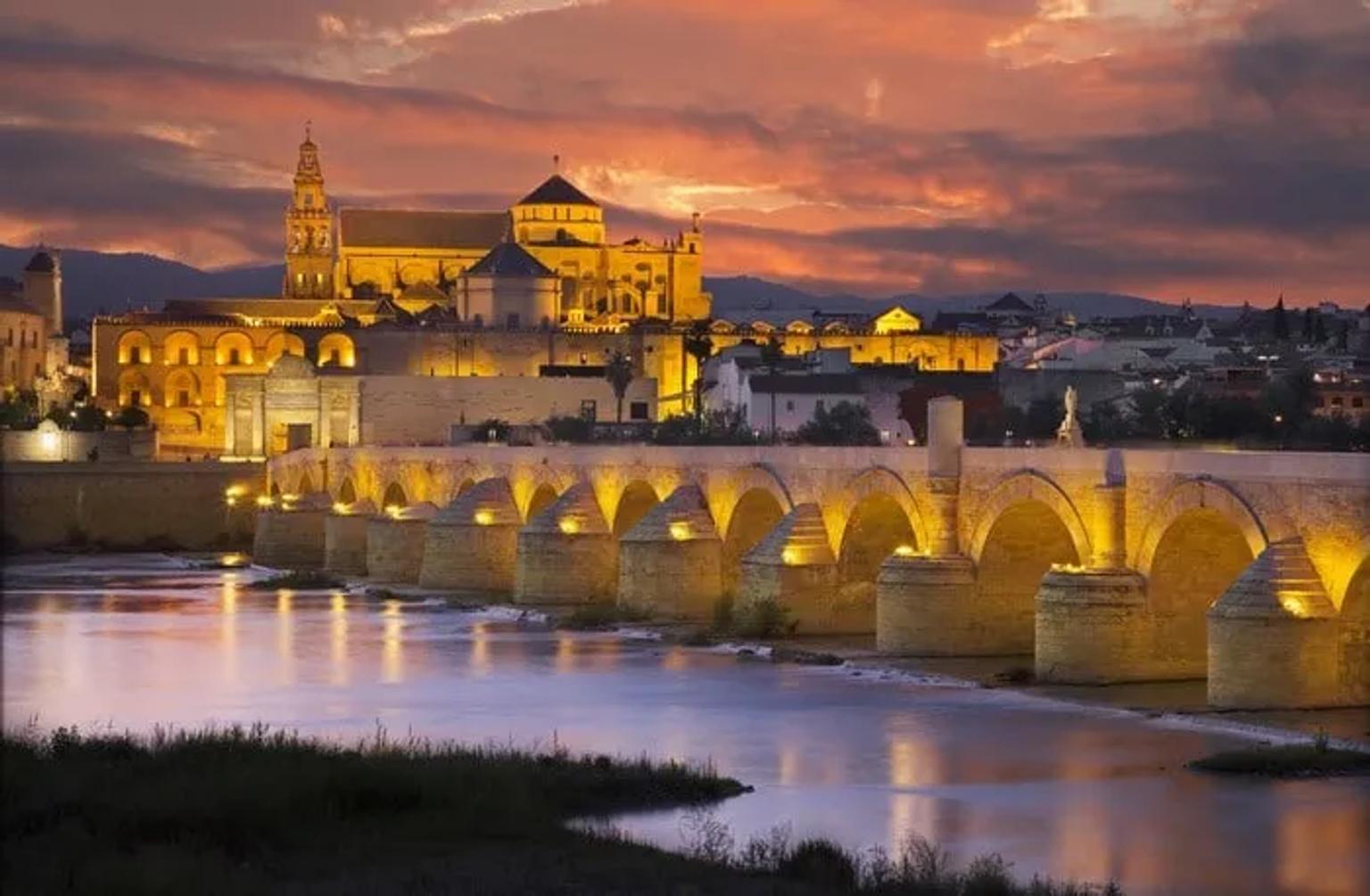 Puente romano iluminado con la ciudad de Córdoba y un atardecer espectacular.