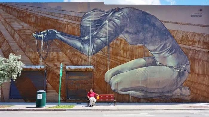 Mural artístico de gran tamaño con figura humana en posición de oración. Persona sentada en banco.
