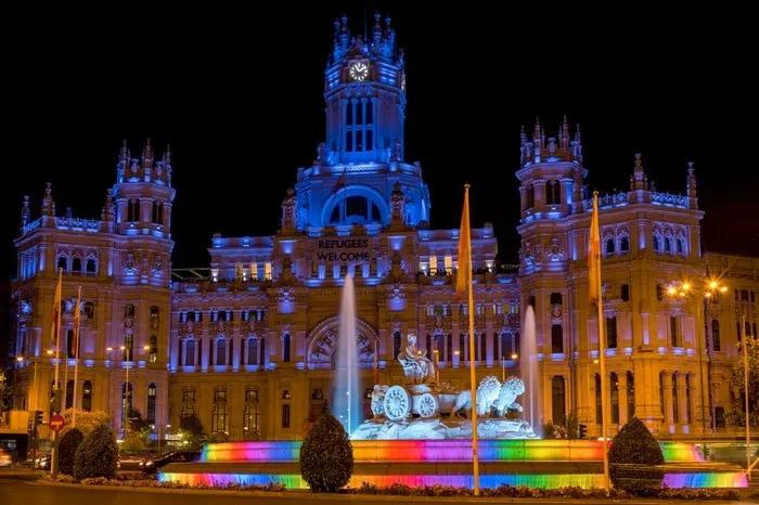 Edificio iluminado en colores, con fuente y mensaje de bienvenida a refugiados.