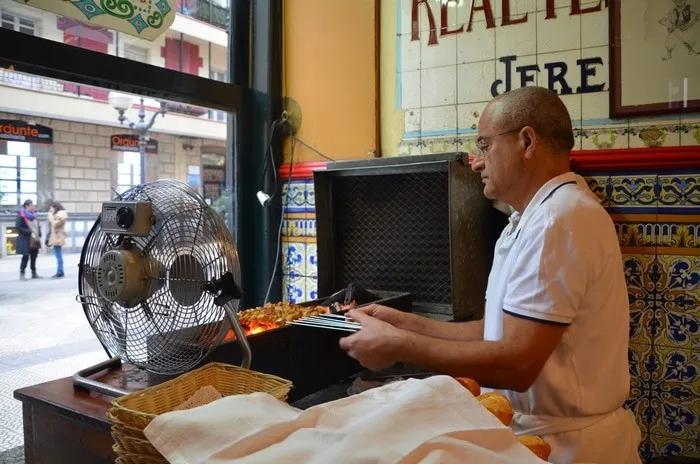 Hombre cocinando pintxos en un bar tradicional de Bilbao. Ventilador y decoración colorida.