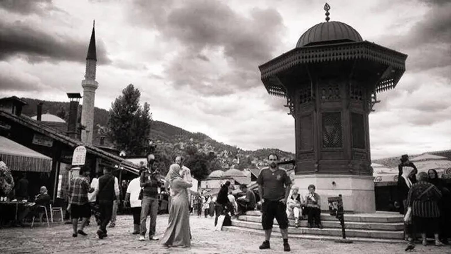 Plaza con un monumento, personas caminando y un ambiente urbano. Nubes en el cielo.