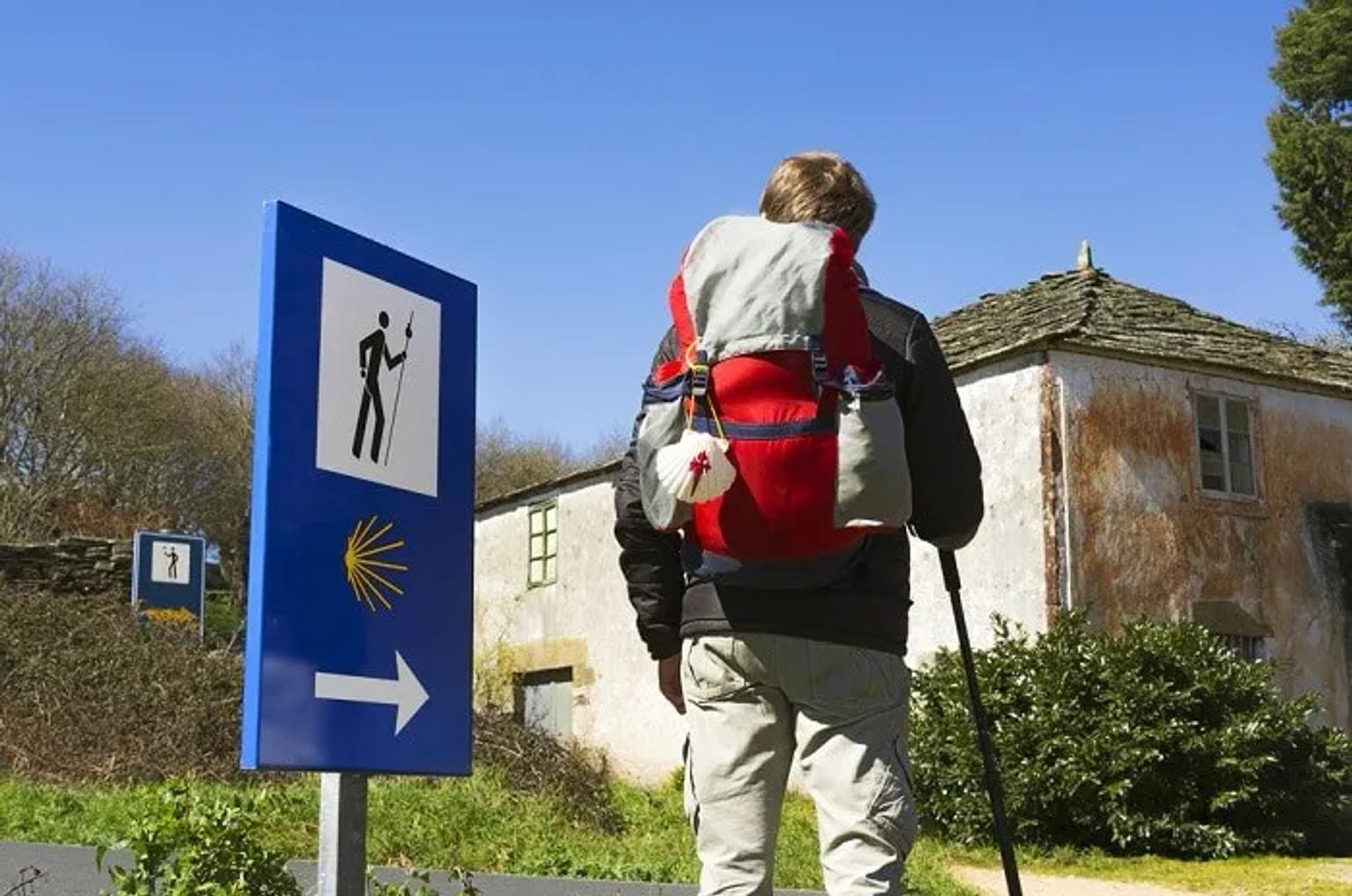 Senderista con mochila frente a señal del Camino de Santiago. Fondo rural y despejado.