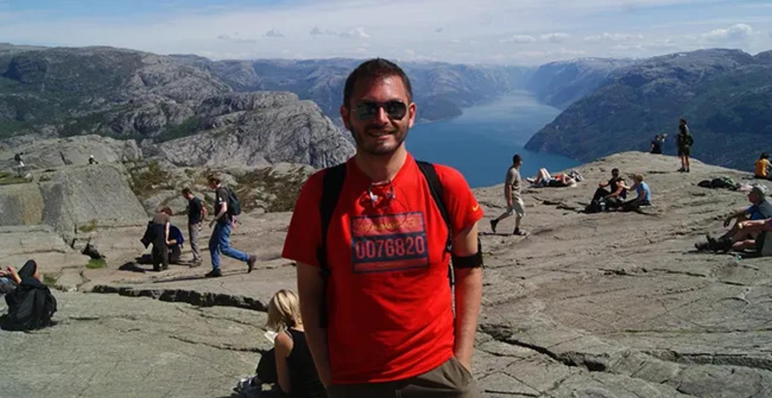 Hombre sonriente en una montaña con paisaje de fiordo al fondo y personas alrededor.