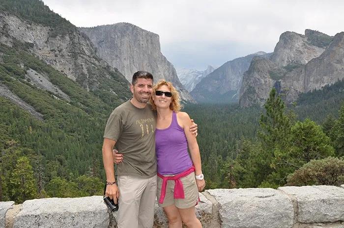 Pareja sonriente en un mirador con montañas y bosque de fondo.