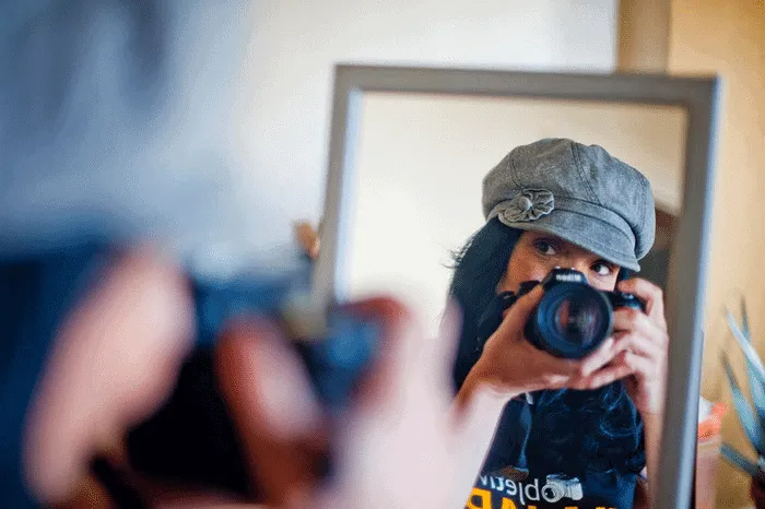 Mujer con gorra se refleja en un espejo mientras sostiene una cámara.