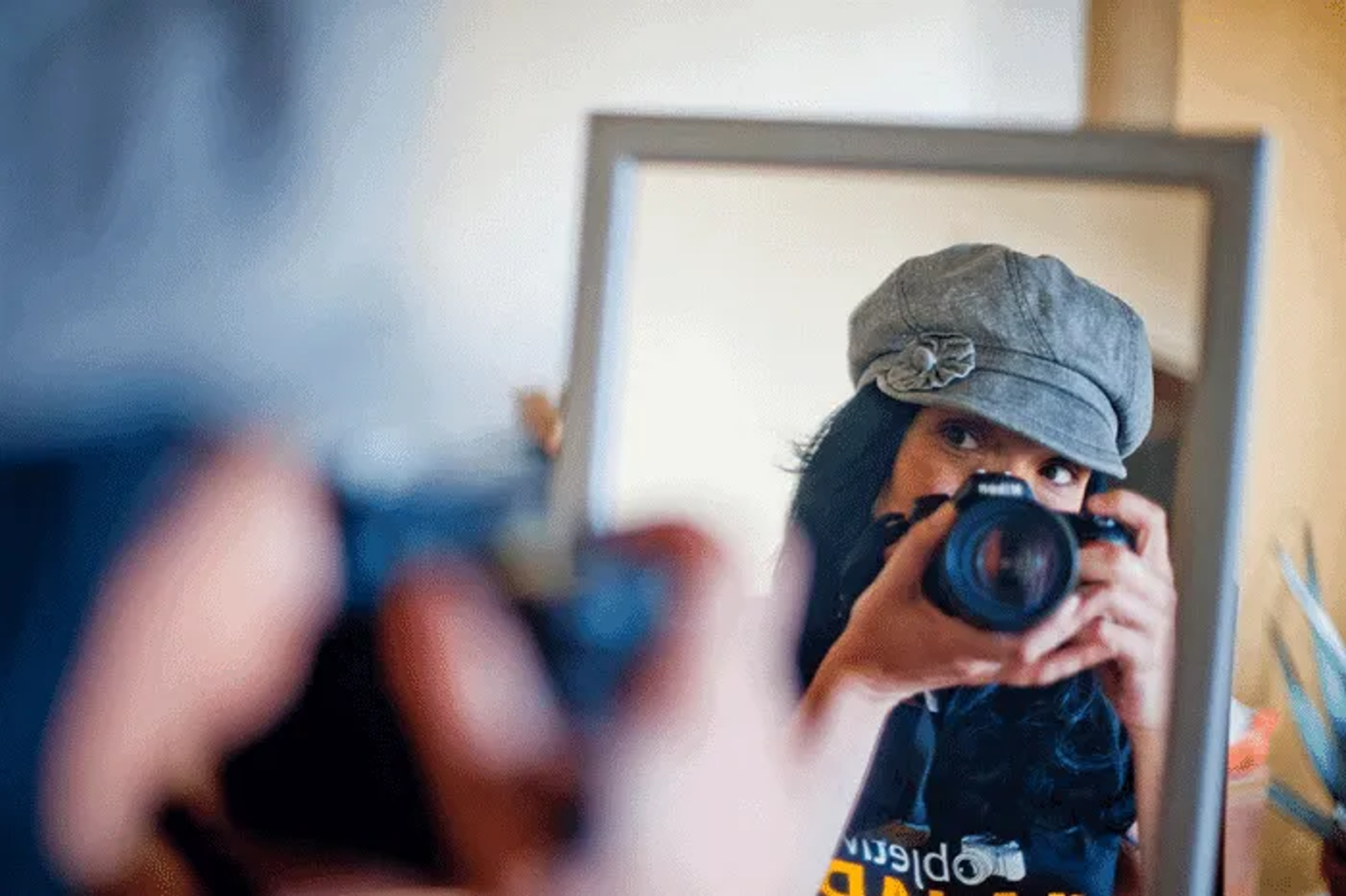 Mujer con gorra se refleja en un espejo mientras sostiene una cámara.
