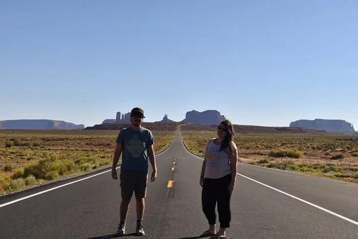 Pareja de viajeros en una carretera desierta con paisajes áridos de fondo.