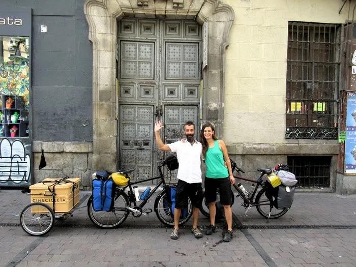 Pareja sonriente junto a bicicletas cargadas, frente a una puerta antigua.