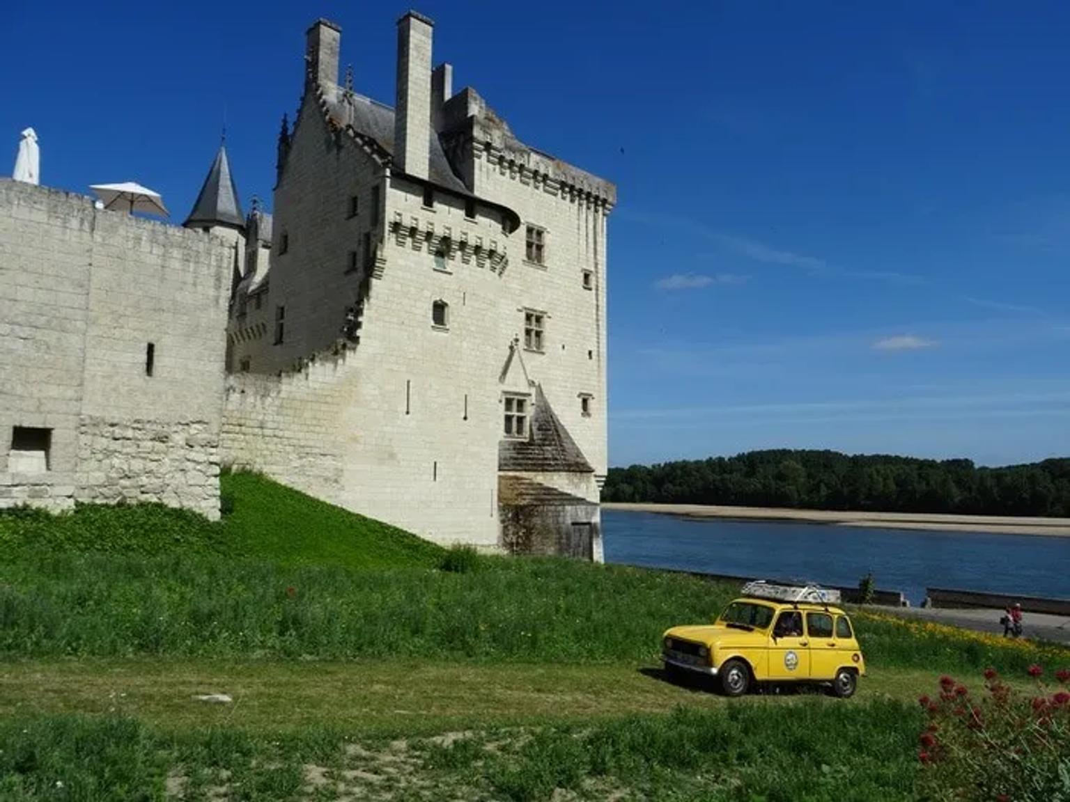 Castillo junto a un río, con un coche amarillo estacionado en el césped.