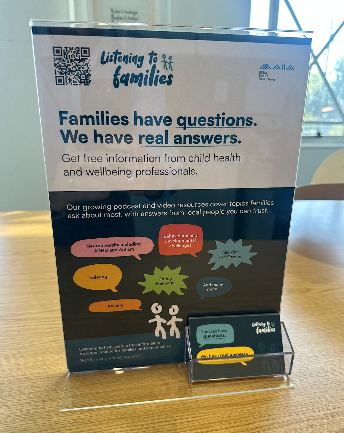 A Listening to Families table talker in a clear acrylic stand on a table, next to a holder of pull-out cards. The sign reads 'Families have questions. We have real answers.'