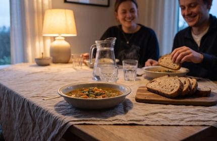 Due scodelle di zuppa, del pane affettato e una caraffa d’acqua su una tavola apparecchiata per due; sullo sfondo due persone sedute a tavola.