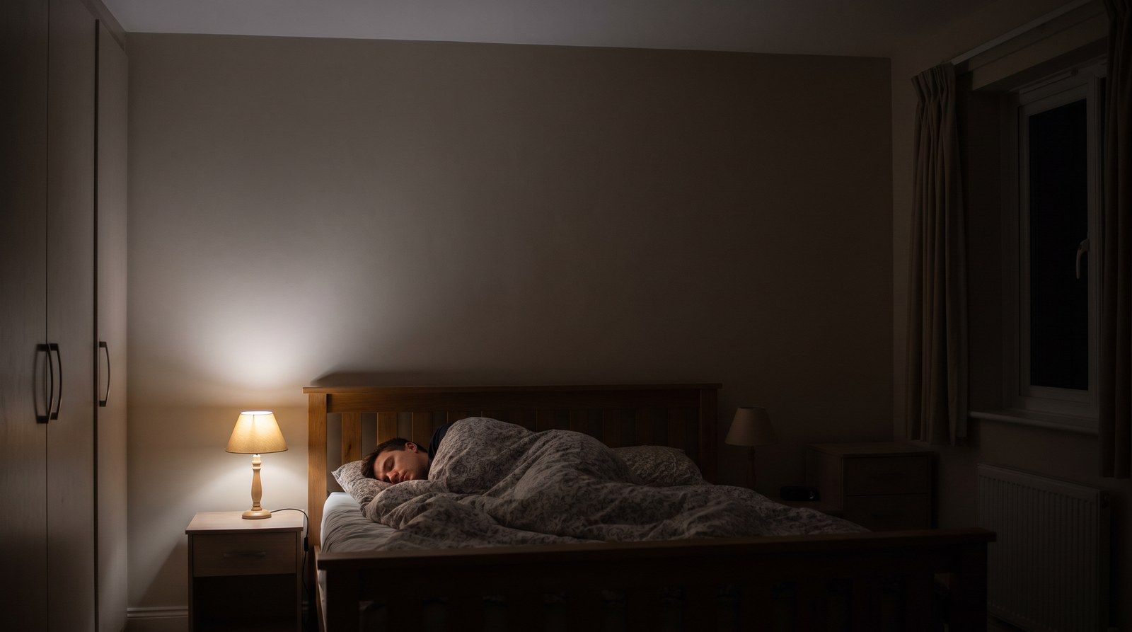 Una persona che dorme nel letto, vista dall’altro lato della stanza, con una piccola lampada ancora accesa.