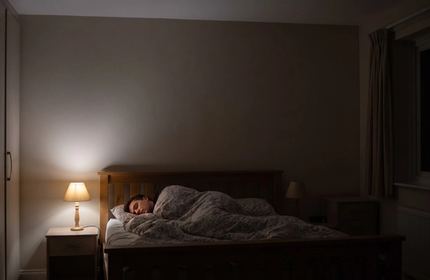 Una persona che dorme nel letto, vista dall’altro lato della stanza, con una piccola lampada ancora accesa.