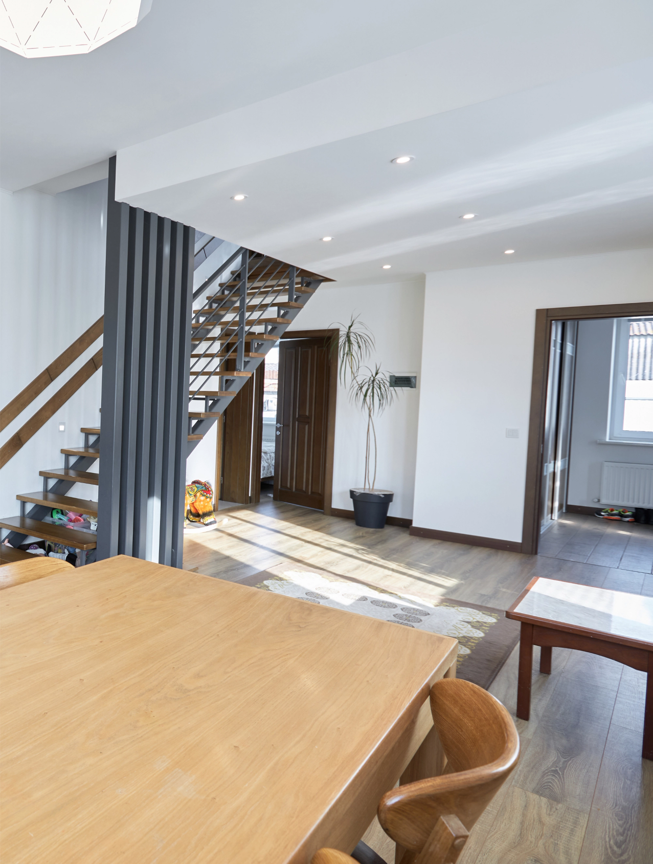 "A modern, open-concept living space with a wooden table, chairs, and a metal staircase. The room is lit by ceiling lights and natural sunlight from large windows.