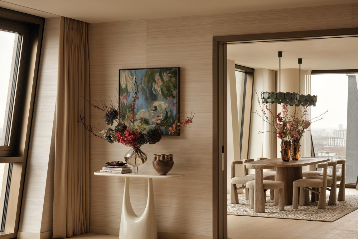 Contemporary apartment interior with textured neutral walls, sculptural console table and floral arrangement, opening into a warm dining room with custom timber table, upholstered chairs and city views.