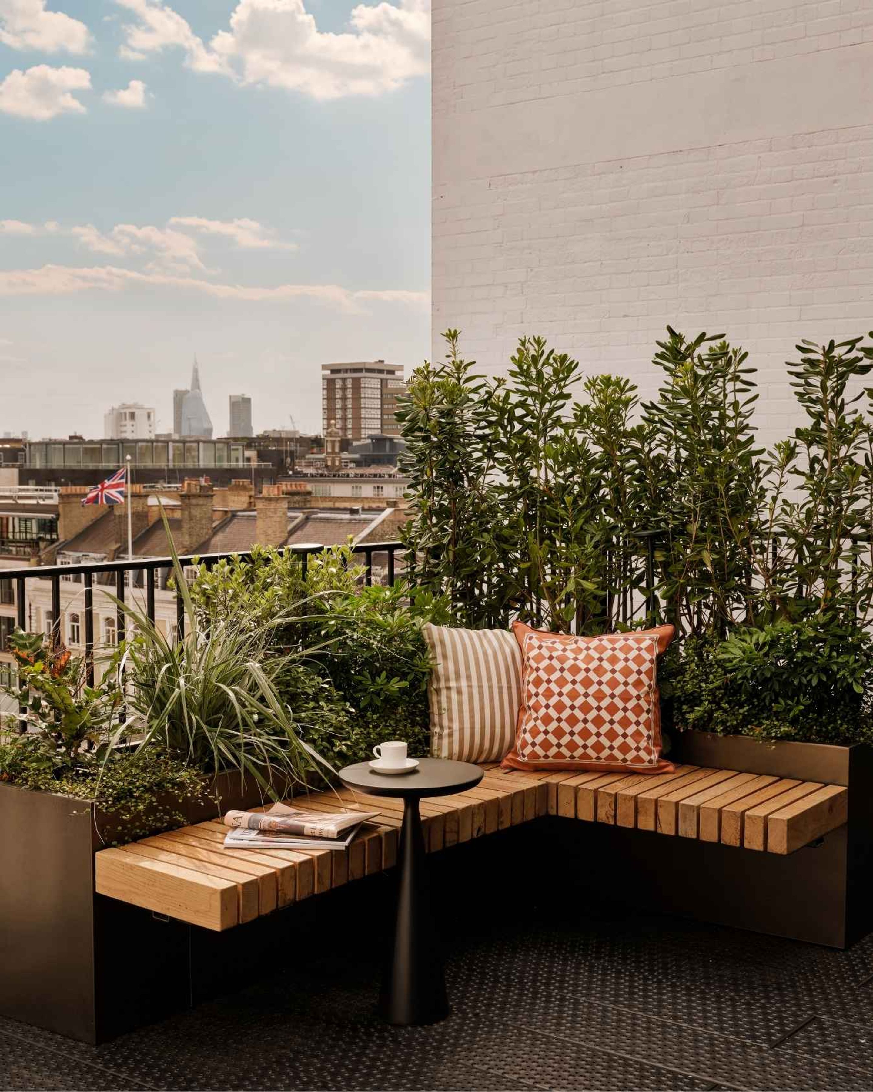 Landscaped London rooftop terrace featuring bespoke wooden seating, patterned cushions, lush planting and city skyline views.