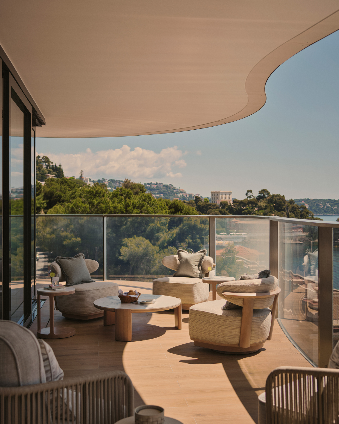 A curved outdoor terrace with low, rounded seating and soft cushions, overlooking a coastal landscape with layered greenery and distant architecture.