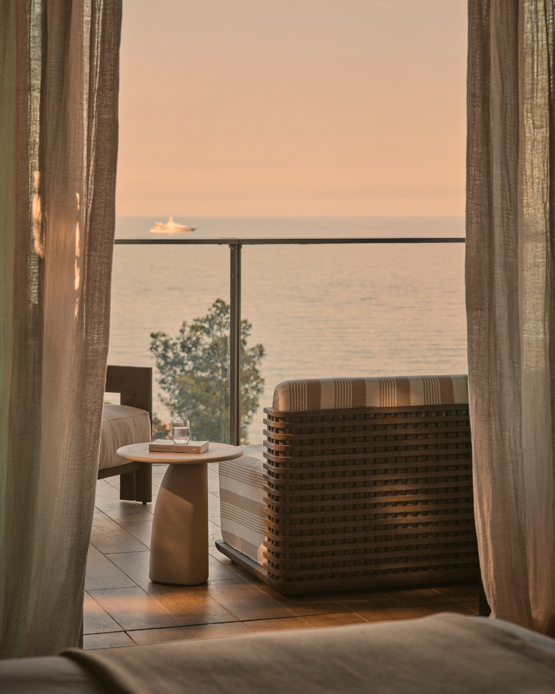 Sea-view terrace with woven outdoor lounge chair, striped cushion and stone side table framed by sheer curtains at sunset, overlooking calm ocean with yacht in the distance.