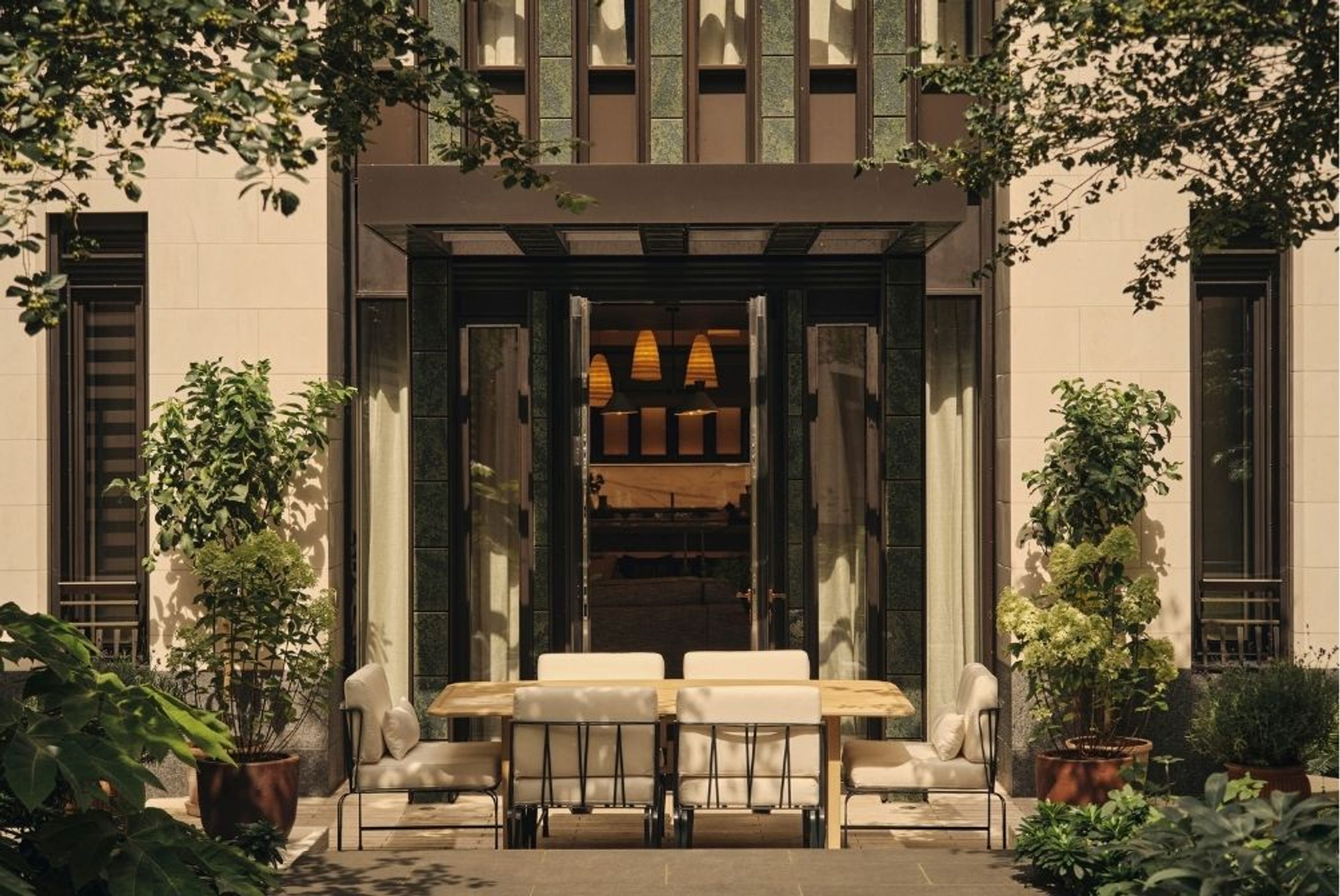 Elegant outdoor dining terrace with a wooden table, upholstered chairs and lush greenery framed by a contemporary stone façade.