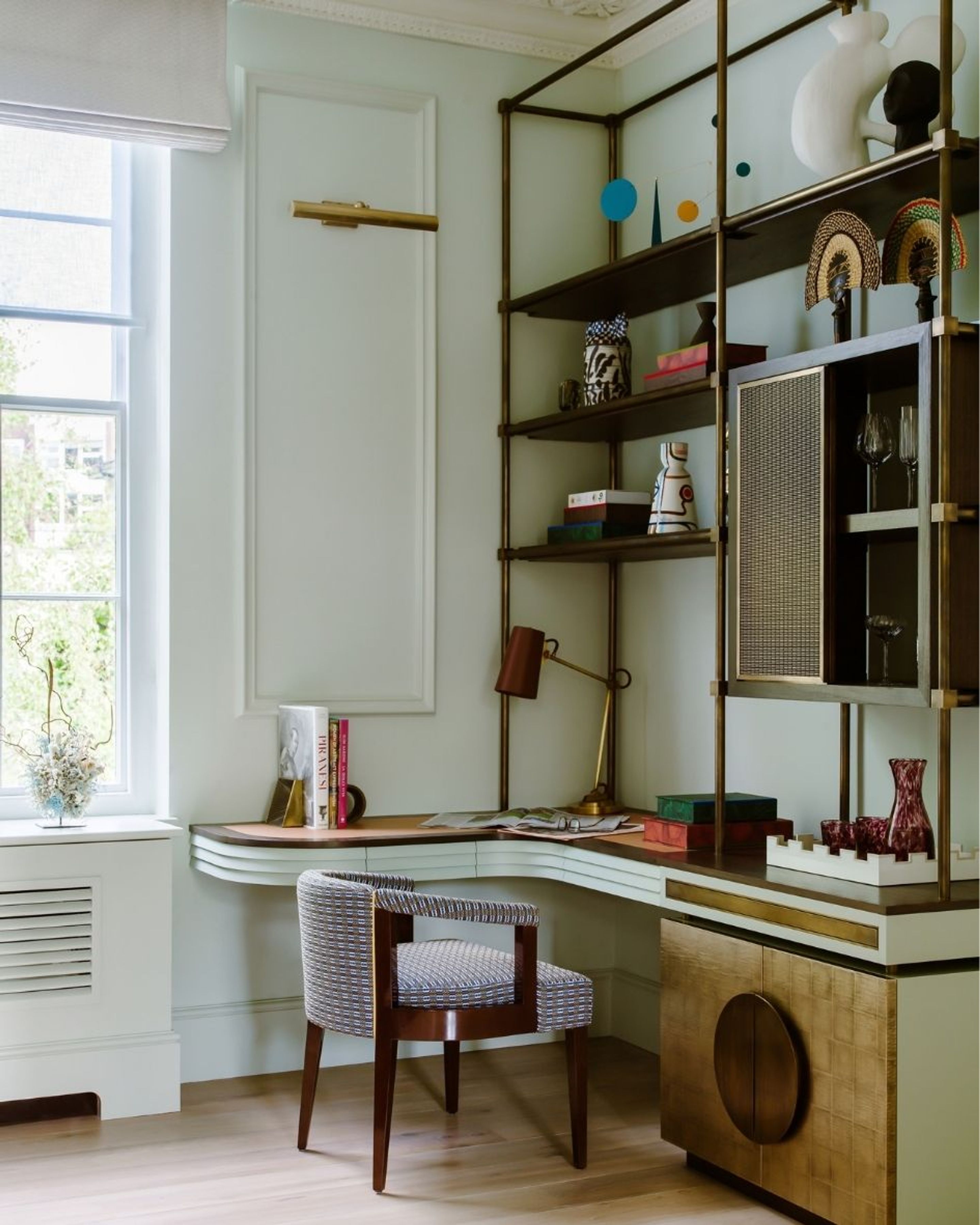 Bespoke home office with curved timber desk, brass-framed shelving, decorative objects and a patterned upholstered chair.