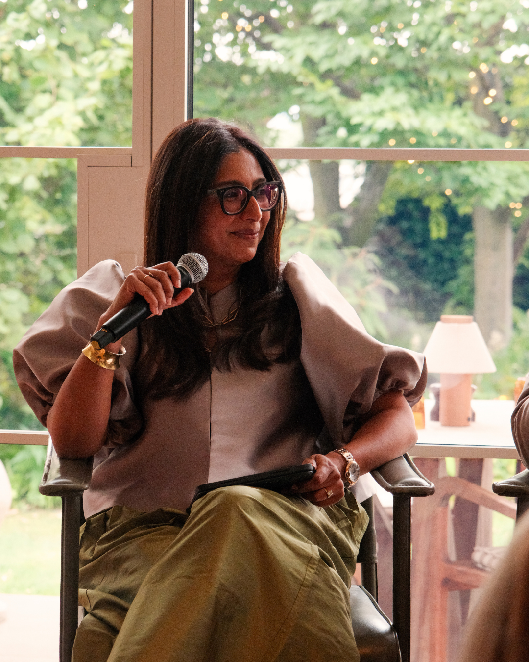 Elicyon Founder and Director Charu Gandhi speaking at an interior design event, seated with a microphone against a leafy garden backdrop.