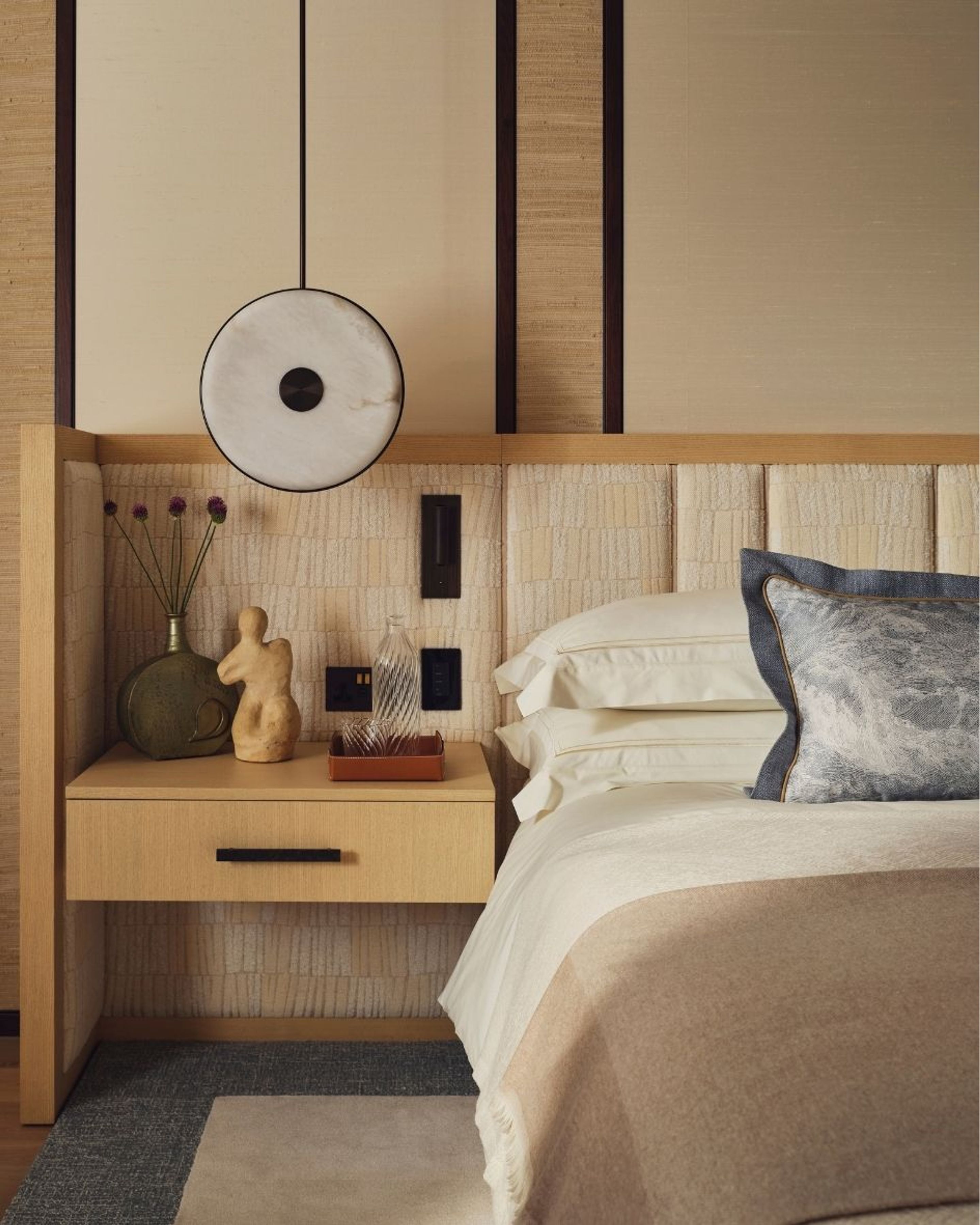 Luxury bedroom interior featuring a bespoke upholstered headboard, layered neutral bedding, timber bedside table and circular pendant light.