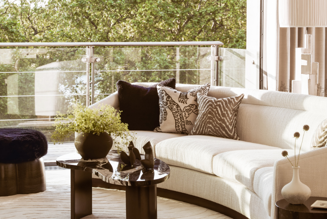 Sunlit living room at One Hyde Park featuring a curved sofa, sculptural furniture, layered textiles and views of mature greenery