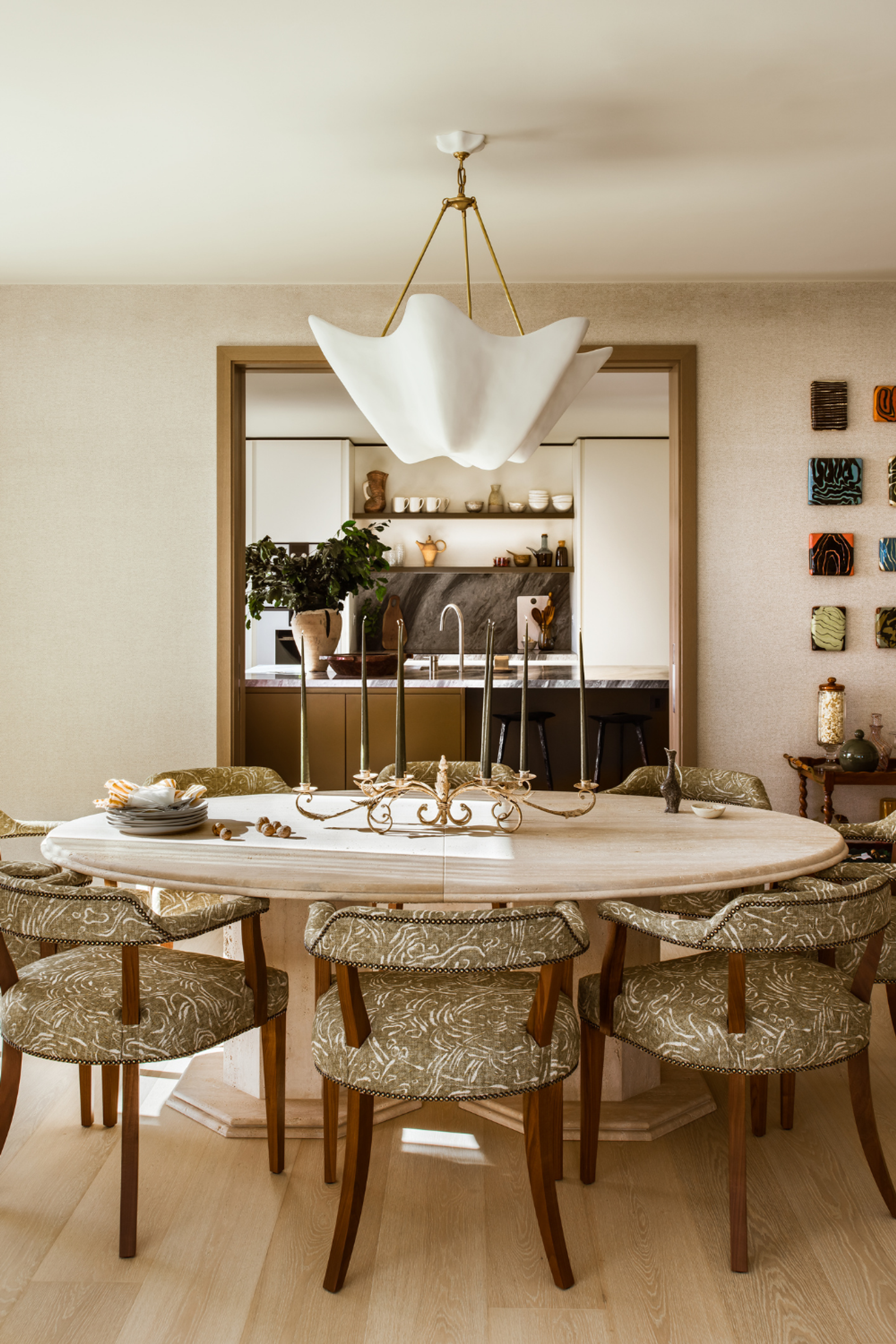 Round stone dining table with upholstered chairs, sculptural pendant lighting and a framed kitchen view in a warm, layered interior.