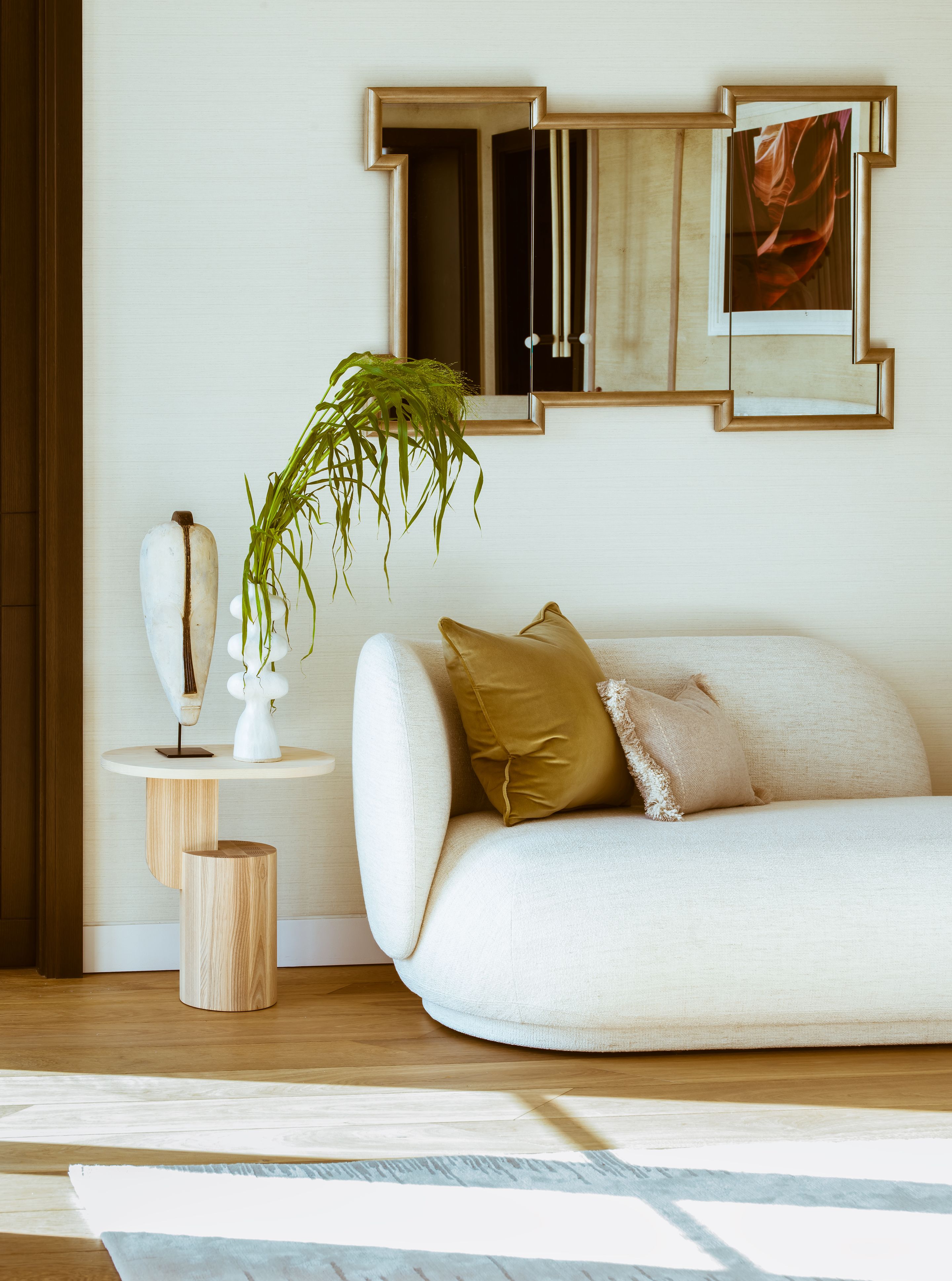 A softly composed seating vignette with a curved upholstered sofa in warm neutral tones, paired with tactile cushions and a sculptural side table. Above, a geometric framed mirror reflects layered textures, while a delicate plant and crafted objects introduce a quiet sense of movement and balance.