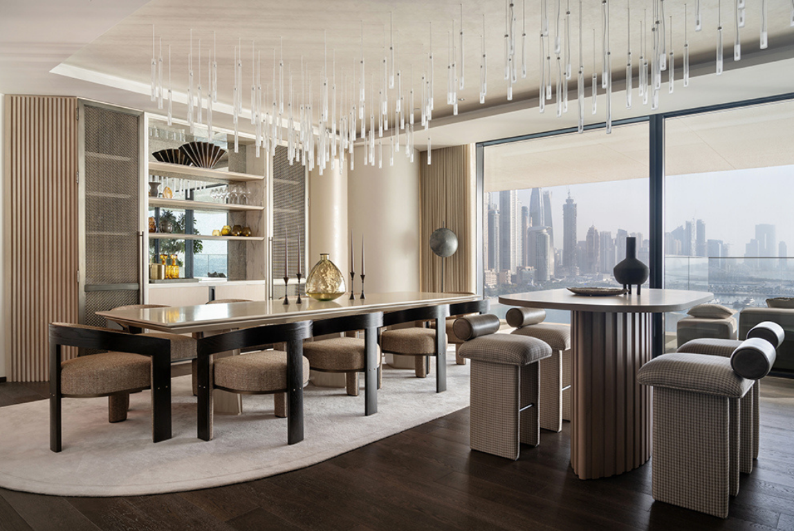 A contemporary dining area with a long timber table, upholstered chairs and suspended glass pendant installation, set against expansive city views.