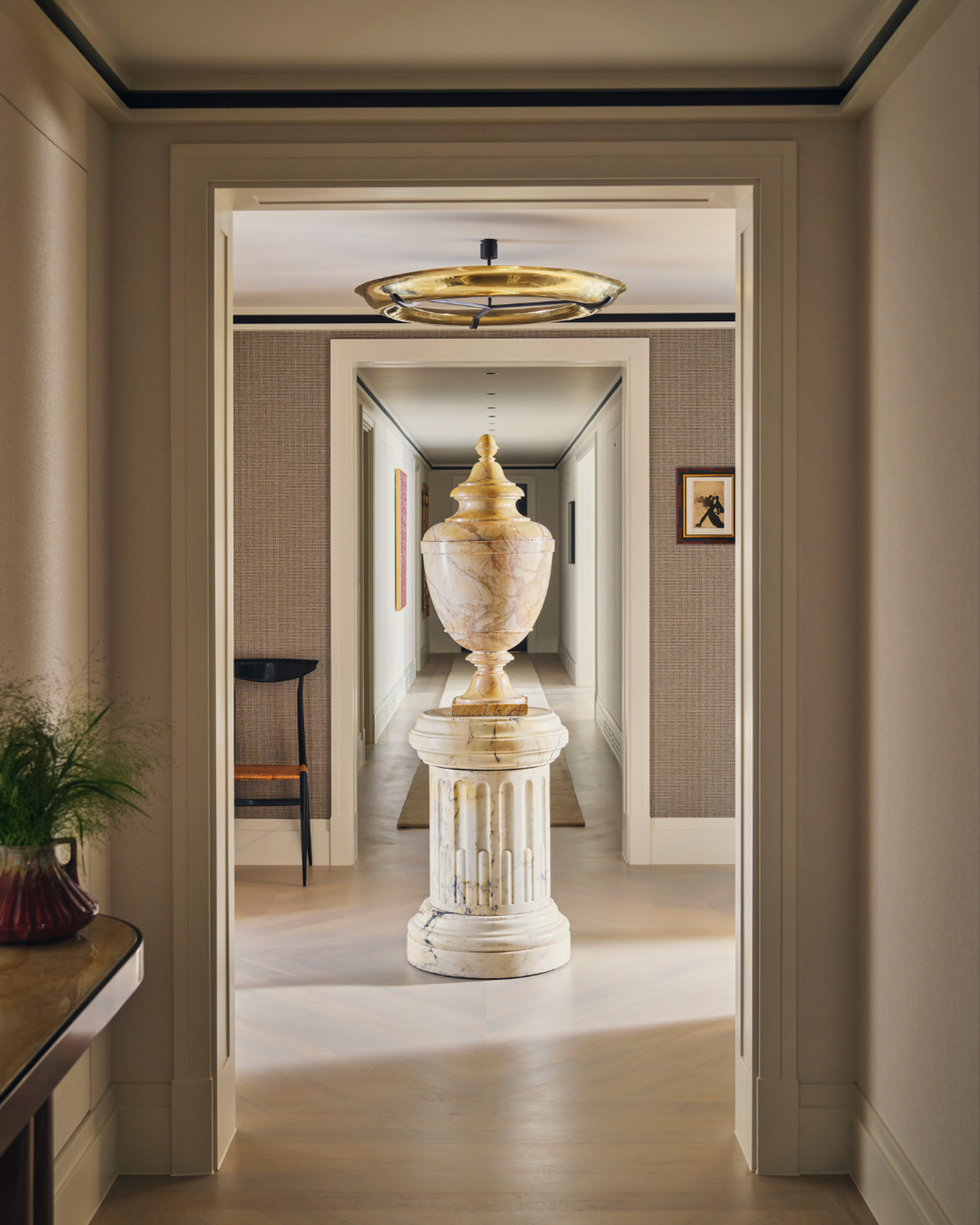 Hallway interior with a classical marble urn sculpture on a pedestal, framed by layered architectural doorways, warm neutral wall finishes and brass ceiling lighting in a refined contemporary residential interior design.