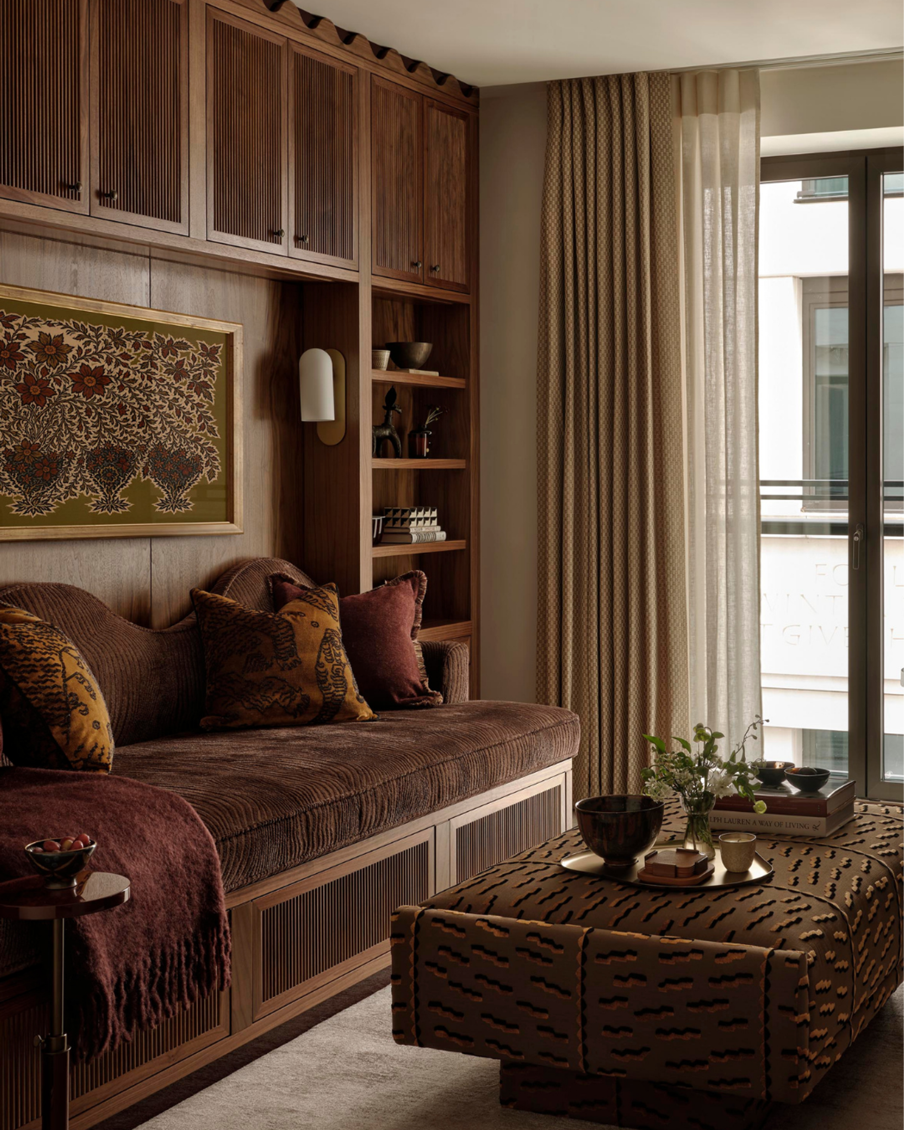 Living room with custom walnut cabinetry, built-in daybed with textured cushions, patterned ottoman coffee table and layered curtains beside a floor-to-ceiling window, showcasing warm contemporary interior design.