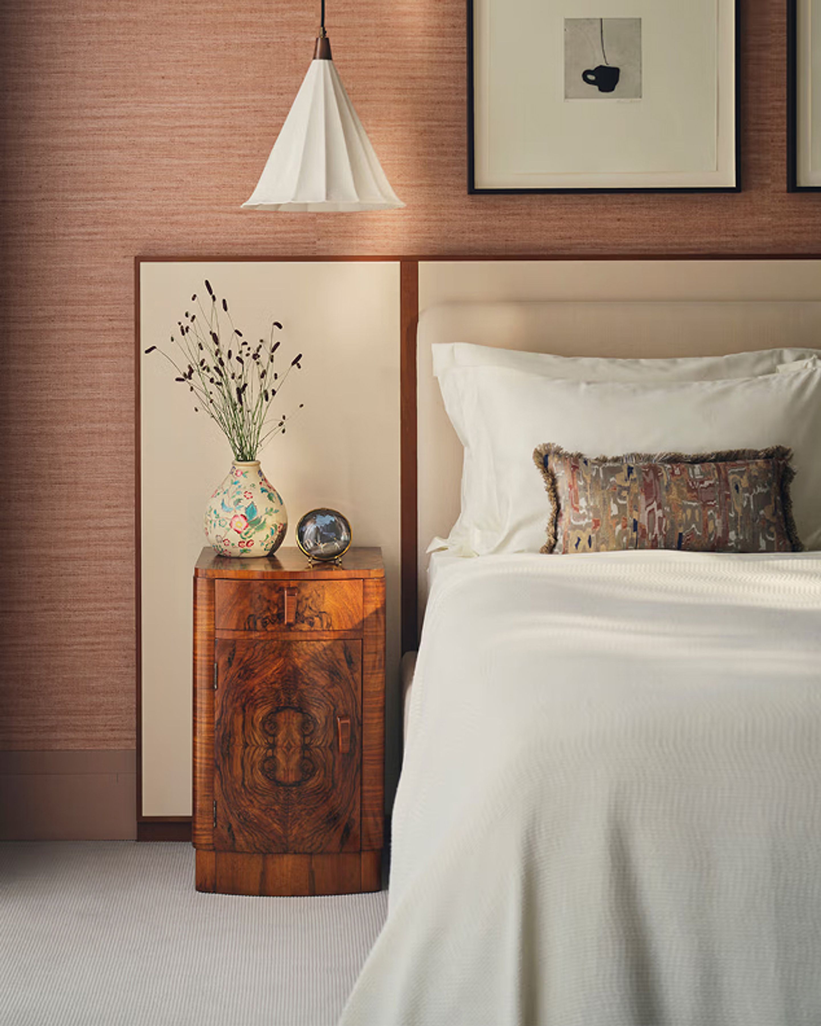 A close up of a bed, side table with a vase, and artwork on the wall.