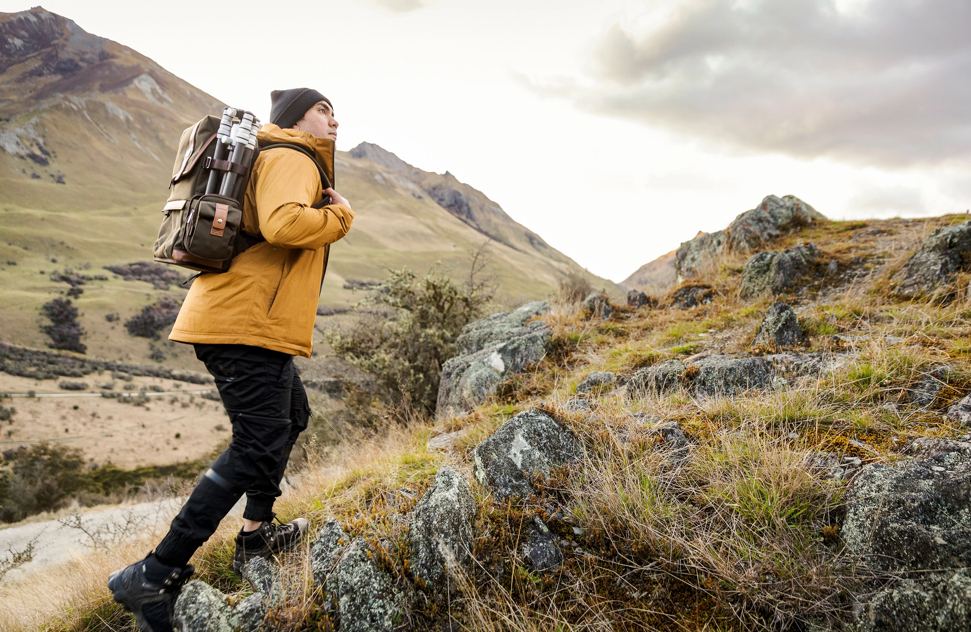 Person wearing a brown outdoor jacket and black beanie, walking in a mountainous landscape. They are carrying a large, rugged backpack equipped with camera tripods on the side, ideal for photography adventures and outdoor explorations.